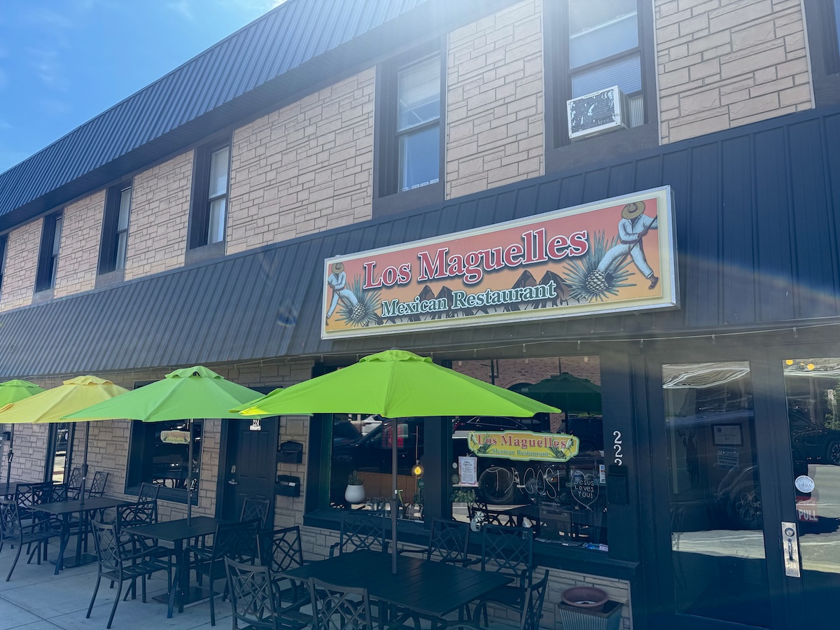 A street-level exterior view of Los Maguelles Mexican Restaurant in Fortville, Indiana. The restaurant is housed in a two-story tan brick building with dark metal siding on the right side. A large rectangular sign above the storefront features the restaurant's name in red cursive and includes illustrations of men harvesting agave plants. On the sidewalk, three bright lime-green patio umbrellas shade black metal tables and chairs, ready for outdoor dining. The glass front windows and a black door with the address "222" are visible beneath a dark grey metal awning.
