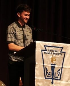 A young man with short dark hair and a beard stands smiling at a podium on a stage with a dark backdrop. He is wearing a short-sleeved, grey and black patterned button-down shirt. The podium is draped with a white banner for the National Honor Society, featuring its official blue and gold torch emblem and the letters "C S L S."