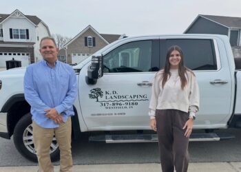 A medium shot of a man and a woman standing in front of a white K.D. Landscaping crew-cab pickup truck. The man on the left wears a blue patterned button-down shirt and khaki pants, and the woman on the right wears a cream-colored sweatshirt and brown joggers. The truck's door prominently displays the company logo—a black tree—along with the text "K. D. LANDSCAPING," the phone number "317-896-9180," and "WESTFIELD, IN." The background features suburban houses and a grey paved street under an overcast sky.