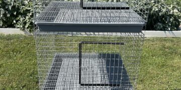 A three-tier, vertical galvanized wire animal cage system stands outdoors on a green lawn. Each level features a front-access door with a black frame and a slide-out grey plastic tray for easy cleaning. A small metal manufacturer's plate is attached to the upper left corner of the top cage. In the background, a lush green field and garden area sit under a clear blue sky.