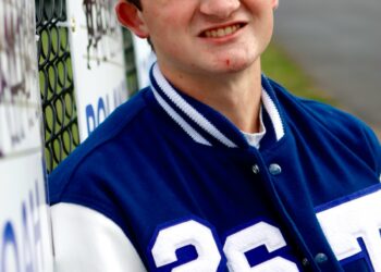 A high-angle portrait of a teenage boy with brown hair and a slight smile. He is leaning against a chain-link fence decorated with team signs. He wears a blue and white varsity jacket featuring the number "26" on the chest. The background shows an out-of-focus athletic field or track area.