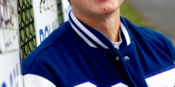 A high-angle portrait of a teenage boy with brown hair and a slight smile. He is leaning against a chain-link fence decorated with team signs. He wears a blue and white varsity jacket featuring the number "26" on the chest. The background shows an out-of-focus athletic field or track area.