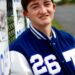 A high-angle portrait of a teenage boy with brown hair and a slight smile. He is leaning against a chain-link fence decorated with team signs. He wears a blue and white varsity jacket featuring the number "26" on the chest. The background shows an out-of-focus athletic field or track area.