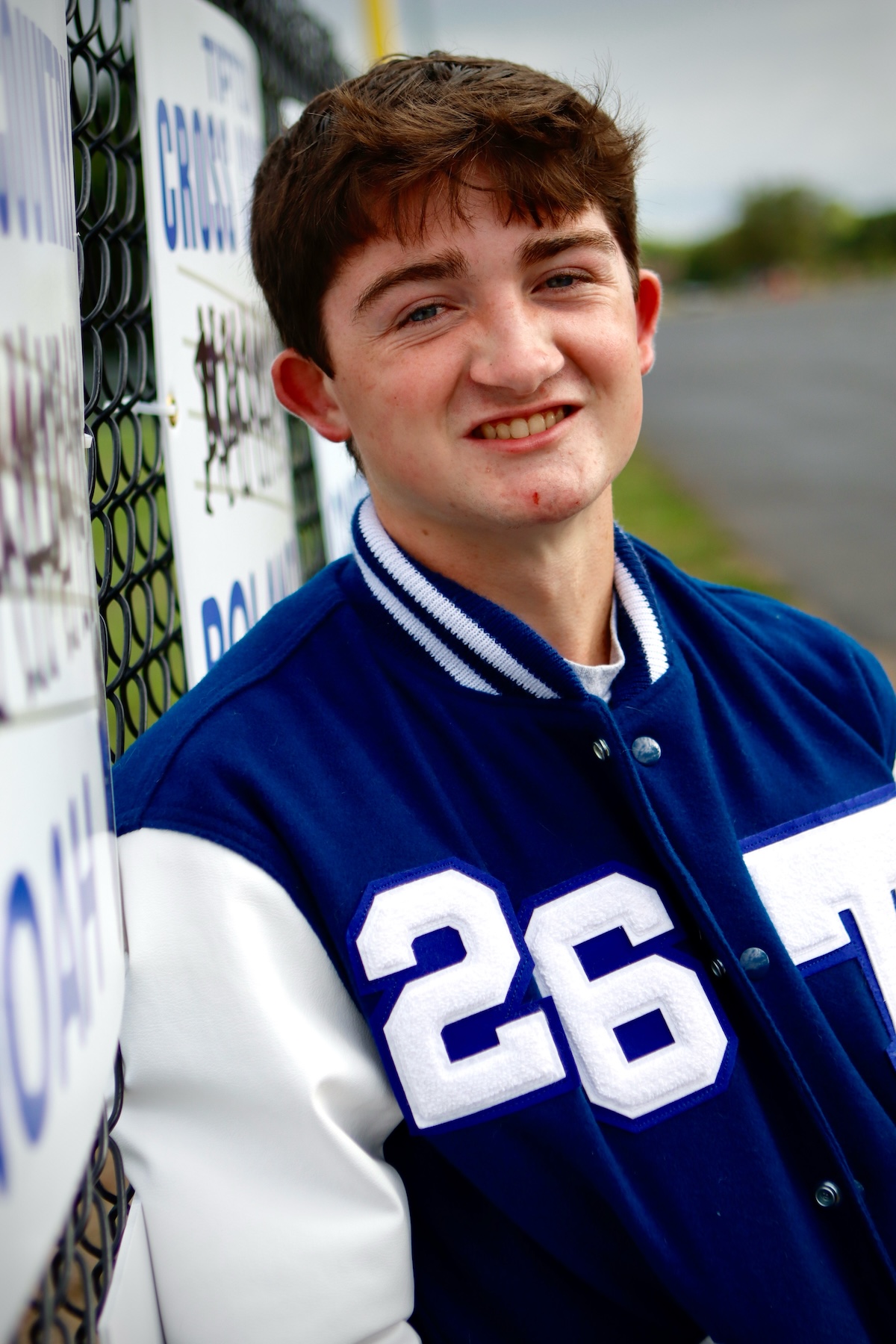 A high-angle portrait of a teenage boy with brown hair and a slight smile. He is leaning against a chain-link fence decorated with team signs. He wears a blue and white varsity jacket featuring the number "26" on the chest. The background shows an out-of-focus athletic field or track area.