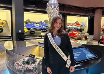 A medium full-shot of a smiling young woman, identified as a 500 Festival Princess for 2026, posing inside the Indianapolis Motor Speedway Museum. She is wearing a black blazer over a light blue blouse, a small sparkling tiara, and a white sash that reads "500 FESTIVAL PRINCESS 2026." She stands in front of a sleek, modern display case illuminated with blue neon lights. Directly behind her is the iconic, towering silver Borg-Warner Trophy, and in the background, several historic Indy 500 race cars, including a blue and yellow #66 Sunoco car, are on display in glass-fronted cases.