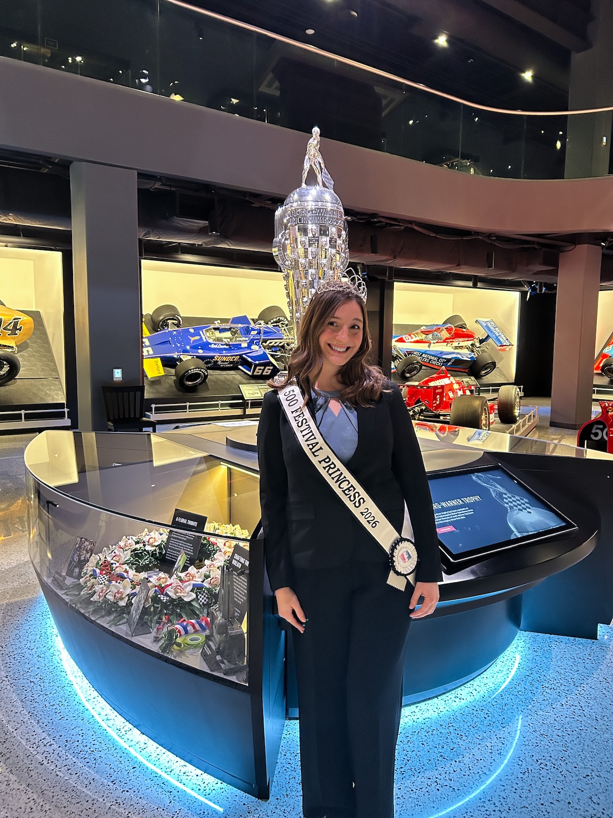 A medium full-shot of a smiling young woman, identified as a 500 Festival Princess for 2026, posing inside the Indianapolis Motor Speedway Museum. She is wearing a black blazer over a light blue blouse, a small sparkling tiara, and a white sash that reads "500 FESTIVAL PRINCESS 2026." She stands in front of a sleek, modern display case illuminated with blue neon lights. Directly behind her is the iconic, towering silver Borg-Warner Trophy, and in the background, several historic Indy 500 race cars, including a blue and yellow #66 Sunoco car, are on display in glass-fronted cases.