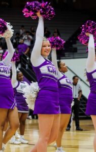 A young cheerleader with blonde hair and a joyful expression is captured mid-cheer during a game. She is wearing a purple and white uniform featuring a block "CH" logo on the chest and the word "CATS" in vertical white letters on the side of her purple skirt. She has one arm raised high, holding a metallic purple pom-pom, while another cheerleader in a similar uniform and a game official in a striped shirt are visible in the background. The scene is set on a polished basketball court with spectators in the stands.