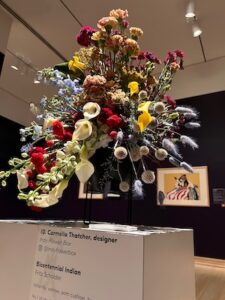 A tall, dramatic floral arrangement by designer Carmella Thatcher of Indy Flower Box at the Saint Louis Art Museum's "Art in Bloom" exhibition. The design features white calla lilies, red carnations, yellow blooms, and tall sprays of dried blue-purple grass. It stands on a white pedestal in front of Fritz Scholder’s painting, "Bicentennial Indian," which is framed on a dark gallery wall in the background.
