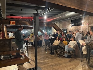 An indoor event at a place like West Sixth Nulu or The Hi-Mark, featuring a man speaking or performing for a seated audience. The room has industrial charm with exposed brick walls, dark wooden ceiling beams, and a polished wood floor. To the left, a man in a dark shirt stands near a microphone and a speaker, looking down at a glass of beer on a wooden counter. The audience is spread throughout the warm, dimly lit space, with some people sitting on folding chairs and others standing in the background near a bar area. A red glowing "EXIT" sign is visible on the left.
