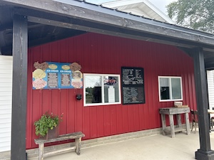 The red-sided exterior of Cowlick's Ice Cream Shoppe's walk-up window area. A colorful "Ice Cream" menu featuring hard-dip and soft-serve options is mounted on the left, next to a service window. A black board with a detailed food menu, including subs and wraps, is positioned between the two windows. Small wooden benches and a rustic table sit on the concrete porch under a dark wooden overhang.