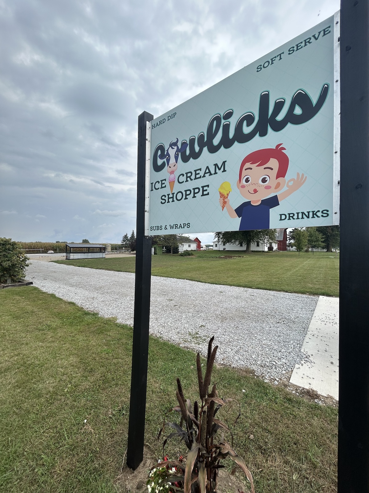 A light blue and white sign for Cowlicks Ice Cream Shoppe stands in front of a green lawn and a gravel driveway. The sign features a cartoon of a young boy with red hair happily holding a yellow ice cream cone. The sign lists offerings such as "Hard Dip," "Soft Serve," "Subs & Wraps," and "Drinks." In the distance, white farm buildings and trees are visible under a cloudy sky.