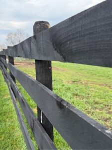 A close-up, perspective shot of a long black wooden fence stretching into the distance. The fence is made of horizontal planks attached to sturdy wooden posts, all painted in a matte black finish. The texture of the wood grain is visible through the paint. Beyond the fence is a lush green pasture under a cloudy sky, with bare trees visible on the horizon.