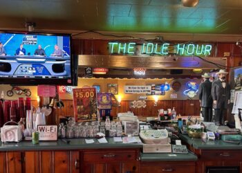 A wide shot of a classic wooden bar counter inside a dimly lit pub. In the center, a bright green neon sign reads "THE IDLE HOUR." To the left, a television displays a "March Madness" sports broadcast. The bar is cluttered with essential items, including stacked glasses, a cash register, napkins, and a "Wi-Fi" sign. Various posters, neon beer signs, and two life-sized statues of men in formal 19th-century attire decorate the back bar area.