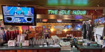 A wide shot of a classic wooden bar counter inside a dimly lit pub. In the center, a bright green neon sign reads "THE IDLE HOUR." To the left, a television displays a "March Madness" sports broadcast. The bar is cluttered with essential items, including stacked glasses, a cash register, napkins, and a "Wi-Fi" sign. Various posters, neon beer signs, and two life-sized statues of men in formal 19th-century attire decorate the back bar area.