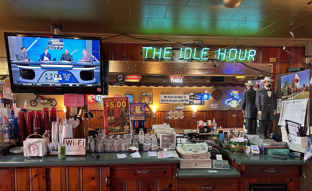 A wide shot of a classic wooden bar counter inside a dimly lit pub. In the center, a bright green neon sign reads "THE IDLE HOUR." To the left, a television displays a "March Madness" sports broadcast. The bar is cluttered with essential items, including stacked glasses, a cash register, napkins, and a "Wi-Fi" sign. Various posters, neon beer signs, and two life-sized statues of men in formal 19th-century attire decorate the back bar area.
