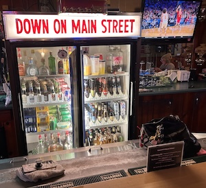 A shot from behind a dark wooden bar counter looking at a double-door reach-in refrigerator. The refrigerator is stocked with various bottled beers and beverages, and it is topped with a bright light box sign that reads "DOWN ON MAIN STREET" in red block letters. To the right, a television mounted on the wall shows a sports game.
