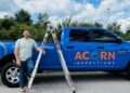 A man in a light green t-shirt and jeans stands smiling next to a bright blue RAM pickup truck. The truck is branded with the orange and white logo for "ACORN INSPECTIONS" and the words "RESIDENTIAL & COMMERCIAL." An aluminum extension ladder is leaning against the side of the truck bed. The scene is outdoors on a sunny day with lush green trees and a blue sky with white clouds in the background.