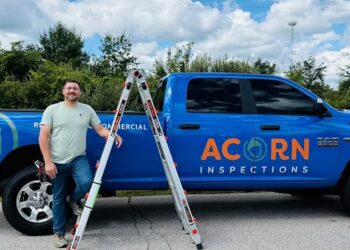 A man in a light green t-shirt and jeans stands smiling next to a bright blue RAM pickup truck. The truck is branded with the orange and white logo for "ACORN INSPECTIONS" and the words "RESIDENTIAL & COMMERCIAL." An aluminum extension ladder is leaning against the side of the truck bed. The scene is outdoors on a sunny day with lush green trees and a blue sky with white clouds in the background.