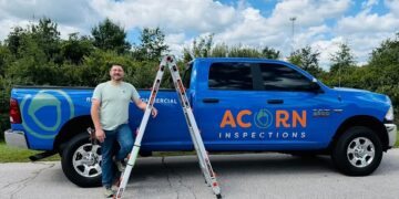 A man in a light green t-shirt and jeans stands smiling next to a bright blue RAM pickup truck. The truck is branded with the orange and white logo for "ACORN INSPECTIONS" and the words "RESIDENTIAL & COMMERCIAL." An aluminum extension ladder is leaning against the side of the truck bed. The scene is outdoors on a sunny day with lush green trees and a blue sky with white clouds in the background.