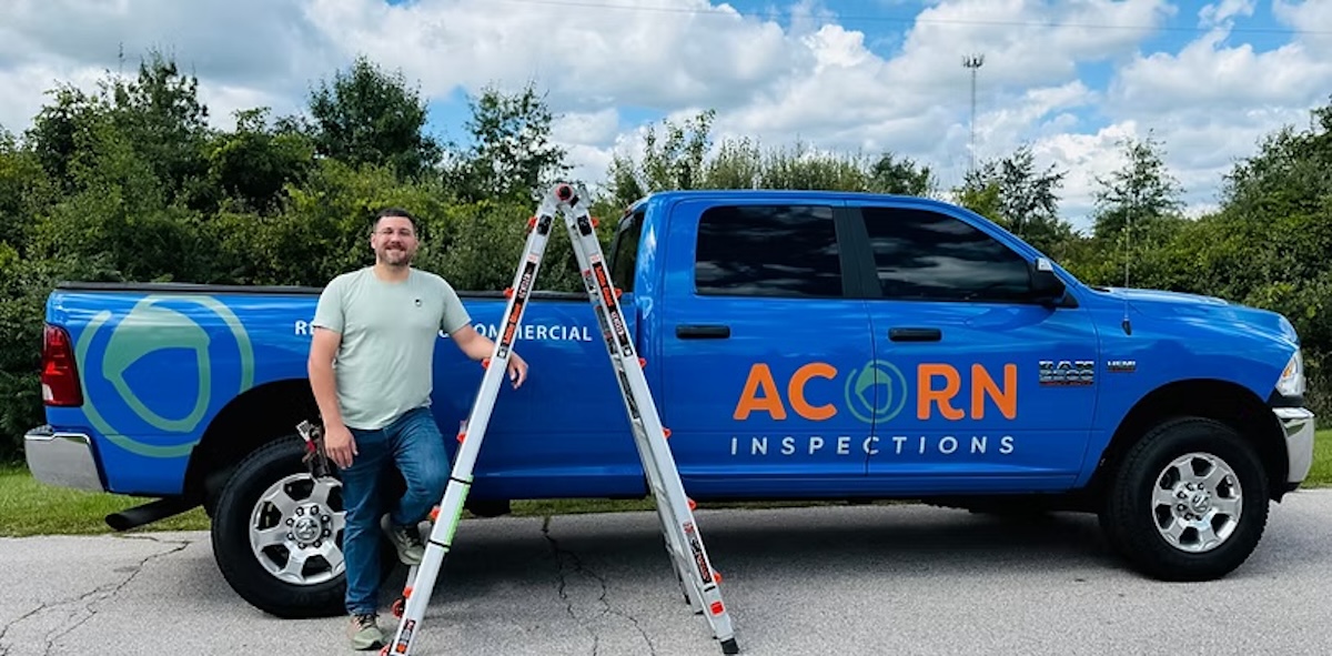 A man in a light green t-shirt and jeans stands smiling next to a bright blue RAM pickup truck. The truck is branded with the orange and white logo for "ACORN INSPECTIONS" and the words "RESIDENTIAL & COMMERCIAL." An aluminum extension ladder is leaning against the side of the truck bed. The scene is outdoors on a sunny day with lush green trees and a blue sky with white clouds in the background.