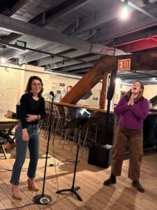 Two women share a joyful moment on a wooden stage in a rustic, dimly lit venue with exposed industrial ceilings and an illuminated "EXIT" sign. The woman on the left, with dark shoulder-length hair and glasses, wears a black long-sleeved top and blue jeans, smiling warmly. To the right, a woman in a vibrant purple sweater and brown textured pants laughs enthusiastically with her hands clasped. A microphone stand and a music rest are positioned in the center between them, and a bar with high stools is visible in the background.