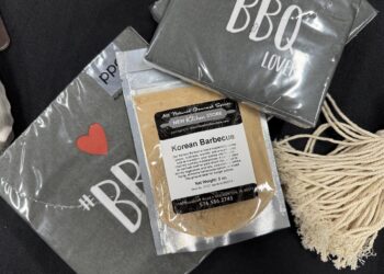 A 2-ounce pouch of "Korean Barbecue" spice blend from the New Kitchen Store sits on a grey t-shirt that reads "#BBQ LOVER" with a red heart. The spice label describes the blend as having "umami, nuttiness, warmth, and a slightly spicy kick." In the background, other packaged grey shirts and a decorative cream-colored tassel are visible on a dark surface.