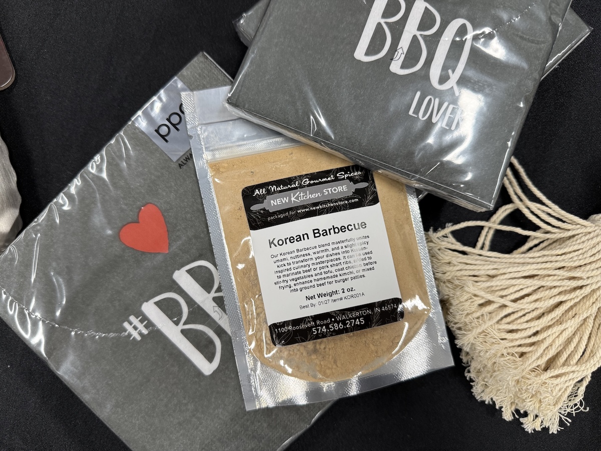 A 2-ounce pouch of "Korean Barbecue" spice blend from the New Kitchen Store sits on a grey t-shirt that reads "#BBQ LOVER" with a red heart. The spice label describes the blend as having "umami, nuttiness, warmth, and a slightly spicy kick." In the background, other packaged grey shirts and a decorative cream-colored tassel are visible on a dark surface.
