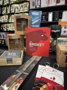 A retail display featuring a variety of grilling and coffee merchandise. In the center is a bright red cookbook titled The Brisket Book by Stephanie Pierson. Surrounding it are several products, including BenShot drinking glasses with embedded bullets, a black Black Rifle Coffee Company mug, a long stainless steel carving knife, and FireWire flexible grilling skewers. In the background, shelves are stocked with more culinary tools and coffee products, creating a one-stop-shop feel for BBQ enthusiasts.