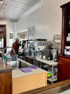 A kitchen area of a cafe featuring stainless steel appliances, including an ice machine and espresso equipment. A person wearing a beanie and a brown shirt is seen in the background working near a window. In the foreground, there is a wooden counter with a sink and purple cleaning cloths, alongside a large, ornate dark wood cabinet.