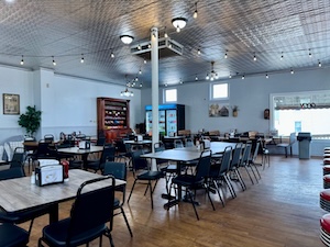 The interior of a spacious diner featuring wooden floors and a pressed-tin ceiling with hanging string lights. Several dark wood tables with black chairs are arranged throughout the room. In the background, there is a large, ornate wooden cabinet, a blue refrigerated display case, and bright windows that let in natural light.