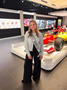 A medium full-shot of a smiling young woman, a 2026 500 Festival Princess, posing inside the modern Indianapolis Motor Speedway Museum. She is wearing a light blue cardigan, black wide-leg trousers, a small sparkling tiara, and a white sash that reads "500 FESTIVAL PRINCESS 2026." Directly behind her is a bright yellow and red #2 Team Penske IndyCar. The background features sleek black walls with illuminated display cases showcasing racing photos and memorabilia under soft, recessed lighting.