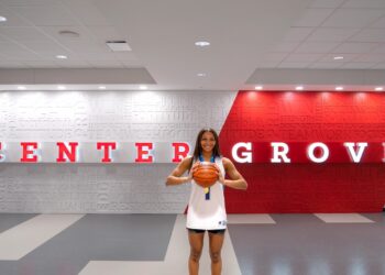 A young female basketball player with long dark hair smiles as she holds a basketball with both hands in front of her. She is wearing a white basketball jersey with blue trim and matching black athletic shorts. She stands in a modern indoor facility against a backdrop that features the words "CENTER GROVE" in large red and white letters. The wall behind her is covered in a word cloud of motivational terms like "TEAMWORK," "FOCUS," and "TOUGHNESS."