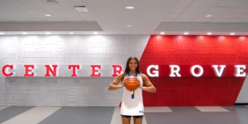 A young female basketball player with long dark hair smiles as she holds a basketball with both hands in front of her. She is wearing a white basketball jersey with blue trim and matching black athletic shorts. She stands in a modern indoor facility against a backdrop that features the words "CENTER GROVE" in large red and white letters. The wall behind her is covered in a word cloud of motivational terms like "TEAMWORK," "FOCUS," and "TOUGHNESS."