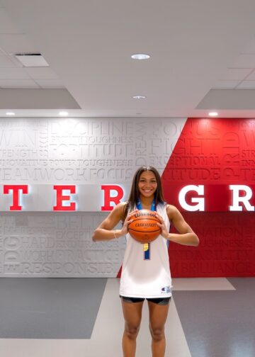 A young female basketball player with long dark hair smiles as she holds a basketball with both hands in front of her. She is wearing a white basketball jersey with blue trim and matching black athletic shorts. She stands in a modern indoor facility against a backdrop that features the words "CENTER GROVE" in large red and white letters. The wall behind her is covered in a word cloud of motivational terms like "TEAMWORK," "FOCUS," and "TOUGHNESS."