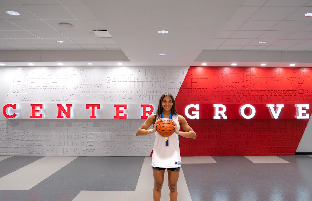 A young female basketball player with long dark hair smiles as she holds a basketball with both hands in front of her. She is wearing a white basketball jersey with blue trim and matching black athletic shorts. She stands in a modern indoor facility against a backdrop that features the words "CENTER GROVE" in large red and white letters. The wall behind her is covered in a word cloud of motivational terms like "TEAMWORK," "FOCUS," and "TOUGHNESS."