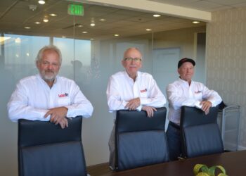 Three men, representing the leadership team of Factory Direct, stand behind a long dark conference table in a modern office. They are all wearing white button-down shirts with the red "Factory Direct" logo on the left chest. The men are leaning their arms on the backs of three black leather executive chairs. The background features a glass-walled conference room with an exit sign and a glimpse of an Indianapolis skyline.