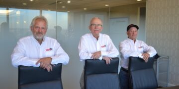 Three men, representing the leadership team of Factory Direct, stand behind a long dark conference table in a modern office. They are all wearing white button-down shirts with the red "Factory Direct" logo on the left chest. The men are leaning their arms on the backs of three black leather executive chairs. The background features a glass-walled conference room with an exit sign and a glimpse of an Indianapolis skyline.