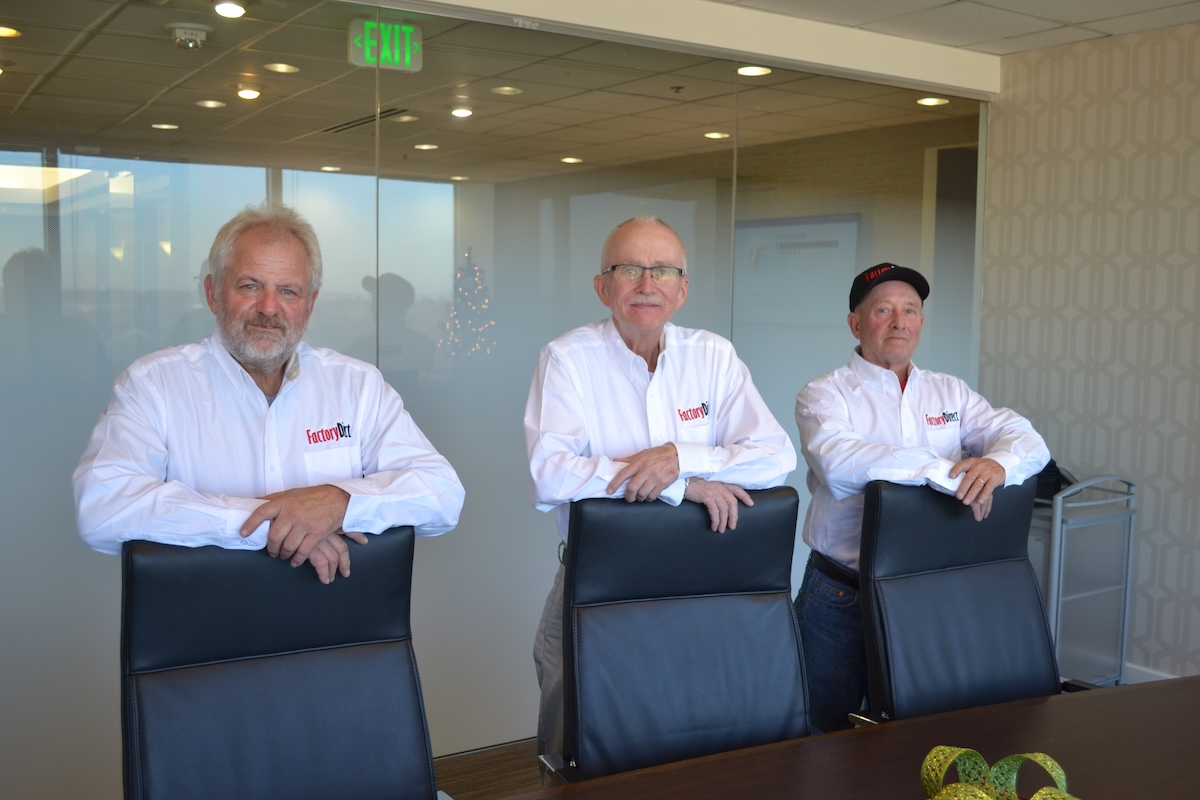 Three men, representing the leadership team of Factory Direct, stand behind a long dark conference table in a modern office. They are all wearing white button-down shirts with the red "Factory Direct" logo on the left chest. The men are leaning their arms on the backs of three black leather executive chairs. The background features a glass-walled conference room with an exit sign and a glimpse of an Indianapolis skyline.