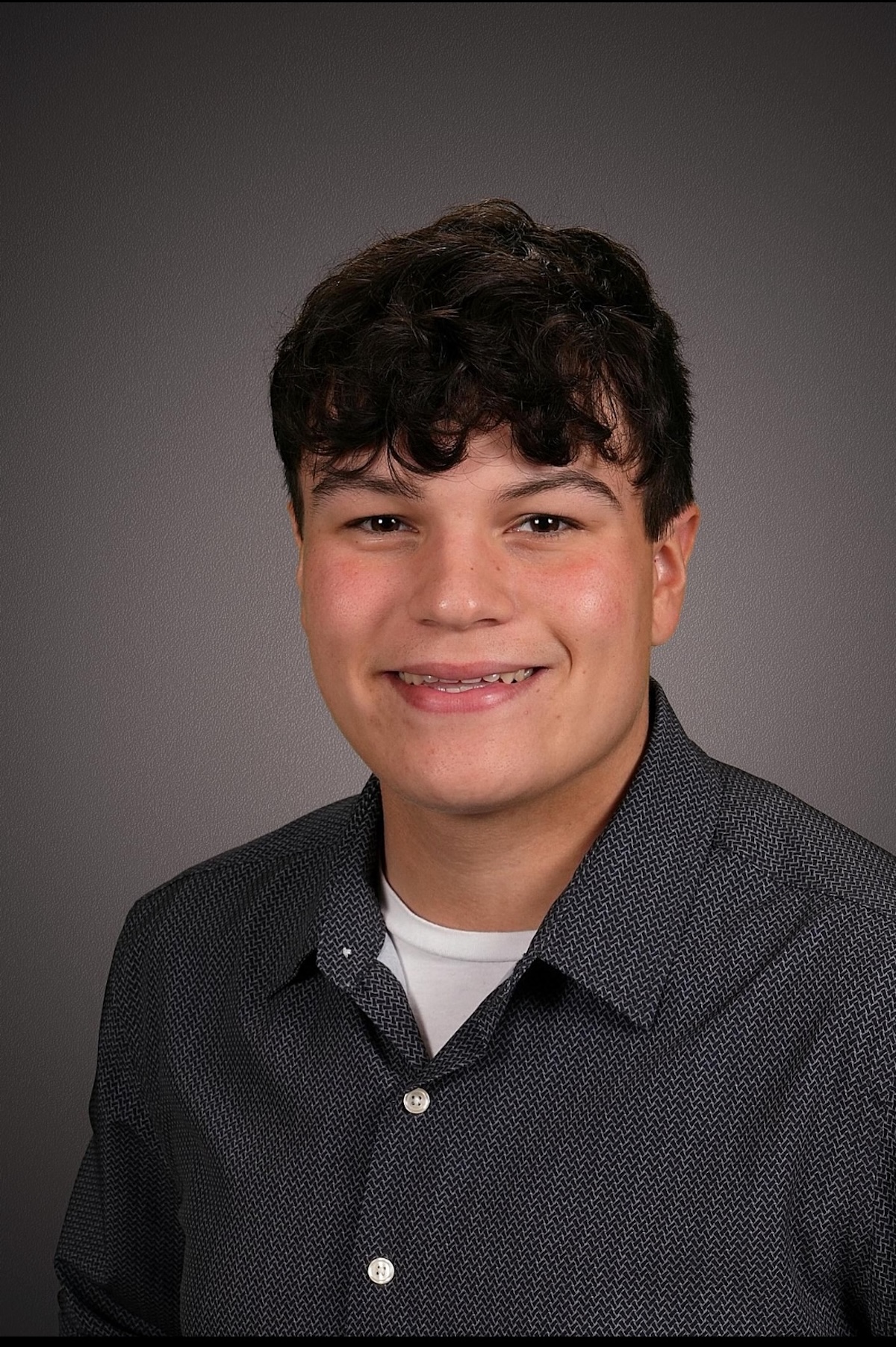 A head-and-shoulders studio portrait of a young man with a warm smile and dark, curly hair. He wears a dark grey button-down shirt with a small geometric pattern over a white t-shirt. The background is a solid, medium-grey gradient.
