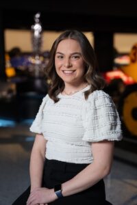 A medium-shot, eye-level portrait of a smiling young woman with shoulder-length, wavy brown hair. She is wearing a white short-sleeved blouse with a textured, layered ruffle pattern and black pants. She is seated with her hands resting in her lap, wearing a black smartwatch with a blue band. The background is a dimly lit, modern interior, likely the Indianapolis Motor Speedway Museum, with blurred warm lights, a blue neon glow, and racing-related exhibits including a large yellow wheel visible on the right.