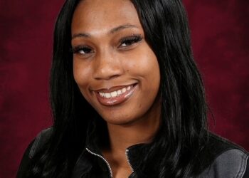 A studio portrait of a young woman with long, dark, wavy hair parted down the middle. She is smiling, showing her teeth, and has a warm expression. She is wearing a black zip-up jacket with grey paneling over a black top. The background is a textured, dark red gradient.