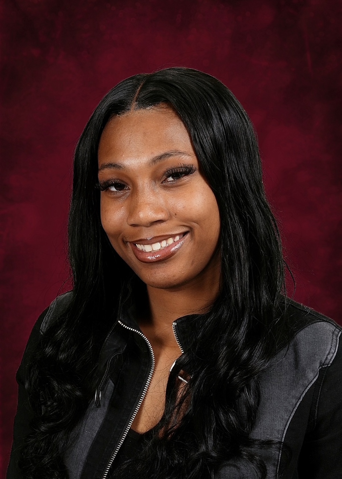 A studio portrait of a young woman with long, dark, wavy hair parted down the middle. She is smiling, showing her teeth, and has a warm expression. She is wearing a black zip-up jacket with grey paneling over a black top. The background is a textured, dark red gradient.