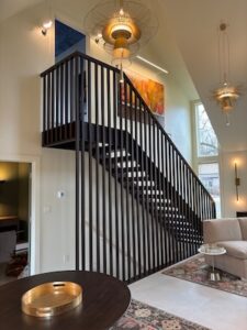 A modern, open-concept living space featuring a striking staircase with a floor-to-ceiling black slat railing. The room has high vaulted ceilings and is illuminated by unique, multi-tiered gold pendant lights. A neutral-toned sofa sits on a patterned area rug in the foreground, and a large piece of vibrant orange abstract art hangs on the wall behind the stairs.