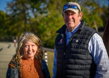 A man and a woman are smiling and posing together outdoors on a sunny day. The man, on the right, wears a tan baseball cap with sunglasses on top, a light blue checkered shirt, and a black quilted vest featuring a red and white logo for "Muncie Central" and a small bear paw emblem. The woman, on the left, has blonde wavy hair and is wearing a rust-colored lace top under a floral cardigan. They are standing near a paved path or bridge with green trees in the soft-focus background.