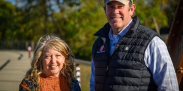 A man and a woman are smiling and posing together outdoors on a sunny day. The man, on the right, wears a tan baseball cap with sunglasses on top, a light blue checkered shirt, and a black quilted vest featuring a red and white logo for "Muncie Central" and a small bear paw emblem. The woman, on the left, has blonde wavy hair and is wearing a rust-colored lace top under a floral cardigan. They are standing near a paved path or bridge with green trees in the soft-focus background.