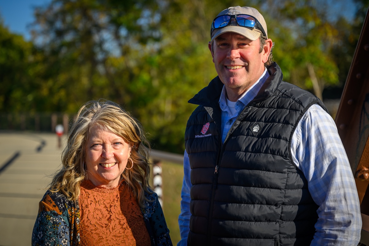 A man and a woman are smiling and posing together outdoors on a sunny day. The man, on the right, wears a tan baseball cap with sunglasses on top, a light blue checkered shirt, and a black quilted vest featuring a red and white logo for "Muncie Central" and a small bear paw emblem. The woman, on the left, has blonde wavy hair and is wearing a rust-colored lace top under a floral cardigan. They are standing near a paved path or bridge with green trees in the soft-focus background.