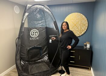 A professional spray tan artist stands next to a large, black Sjolie pop-up tanning tent in a modern salon studio. She is smiling and dressed in all-black attire, holding a professional spray tan gun connected to a machine. The room features a deep teal accent wall decorated with a large, golden sunburst wall sculpture. To the right, a black console table with gold legs holds glass jars filled with white cotton supplies. The Sjolie tent is spacious, featuring clear plastic panels at the top for optimal lighting during sessions.