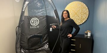 A professional spray tan artist stands next to a large, black Sjolie pop-up tanning tent in a modern salon studio. She is smiling and dressed in all-black attire, holding a professional spray tan gun connected to a machine. The room features a deep teal accent wall decorated with a large, golden sunburst wall sculpture. To the right, a black console table with gold legs holds glass jars filled with white cotton supplies. The Sjolie tent is spacious, featuring clear plastic panels at the top for optimal lighting during sessions.