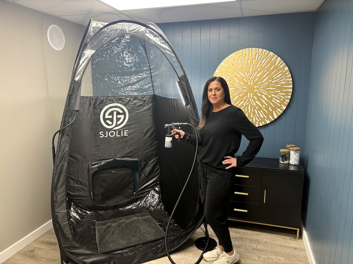 A professional spray tan artist stands next to a large, black Sjolie pop-up tanning tent in a modern salon studio. She is smiling and dressed in all-black attire, holding a professional spray tan gun connected to a machine. The room features a deep teal accent wall decorated with a large, golden sunburst wall sculpture. To the right, a black console table with gold legs holds glass jars filled with white cotton supplies. The Sjolie tent is spacious, featuring clear plastic panels at the top for optimal lighting during sessions.