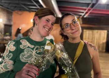 Two women stand together indoors, smiling and posing for a photo. The woman on the left has shoulder-length brown hair and is wearing a green lace blouse and gold earrings. The woman on the right has her dark hair tied back, wears yellow-rimmed glasses, and has a tattoo on her left shoulder. They are both holding small bouquets of white baby’s breath wrapped in clear plastic. The background is a dimly lit room with industrial-style ceilings.